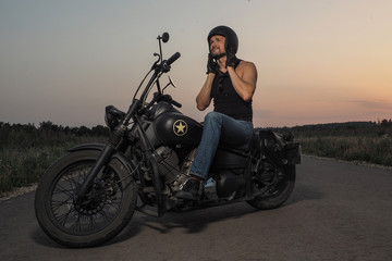 Obraz na płótnie Canvas Masculine motorcycle rider sits on old style bobber, puts on a black helmet, wears blue jeans and black tee on empty road in sunset time 