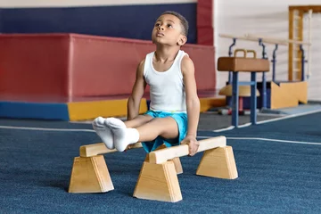 Tuinposter Picture of adorable dark skinned little gymnast competing on parallel bars. Hardworking talented African child exercising at gym, doing acrobatic moves, demonstrating strength, agility and flexibility © Anatoliy Karlyuk
