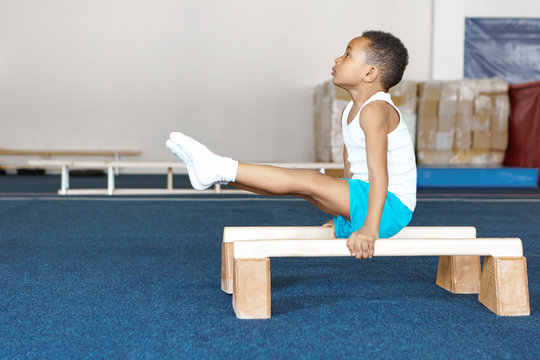 Side view of strong flexible dark skinned male child in sportswear doing exercises on wooden parallel bars, balancing body upright with arms straight, raising legs. Strength, flexibility and endurance