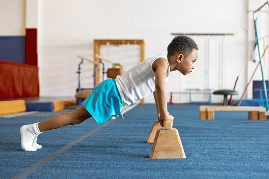 Sideways Shot Of Concentrated Serious Afro American Kid In Sports Clothes Keeping Feet On Floor And Hands On Wooden Bar, Doing Push Ups. Strong Self Determined Black Boy Planking At Fitness Center