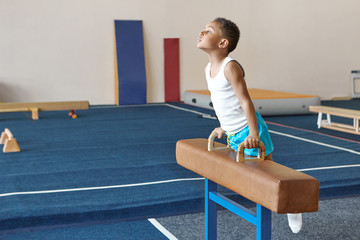 Talented hardworking African boy training indoors at gym, balancing on pommel horse, working on...