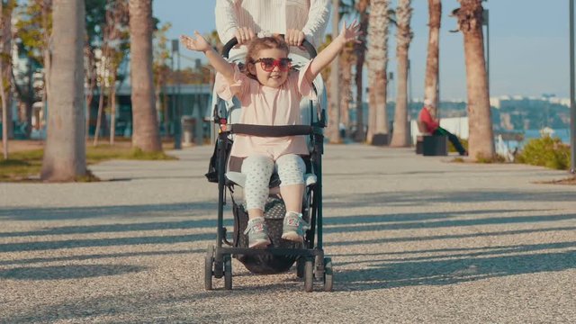 Woman pushes her cute child in a baby stroller down the alley. Girl smiles, waving her hands. Family bonds, cheerful mood, summertime. Happy childhood. Family portrait. Slow motion