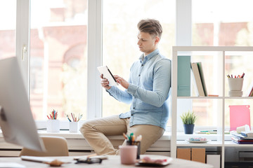 Serious creative young man with hairstyle sitting on window-sill in modern office and viewing online sketches on tablet