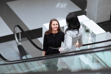 two smiling business women standing on an escalator in a business center