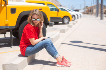 Beautiful woman wearing t-shirt, blue jeans and sunglasses sitting on the pedestrian area near yellow car