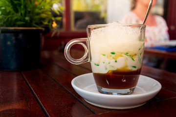 Closeup view, Typical cold sweet South East Asian desert for summer hot season called Lot Chong, Chendol, Dawet, Cendol or Tjendol, served in glass cup on wooden table in Asian restaurant or cafe. 