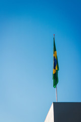 Brazilian flag on pole without wind in front of blue sky.