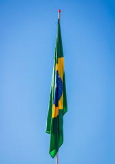 Brazilian flag on pole without wind in front of blue sky.