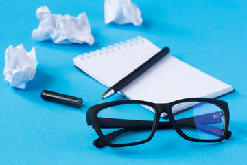 A blank sheet of notepad, pen, glasses and crumpled sheets of paper on a blue background.