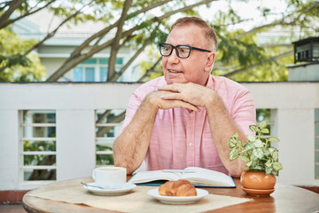 Fototapeta na wymiar Positive pensive senior man enjoying tasty breakfast and good book in outdoor cafe
