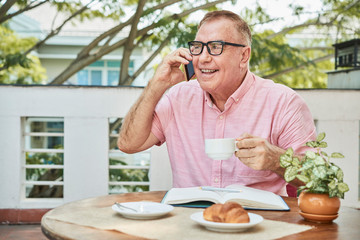 Portrait of happy senior man drinking morning coffee in cafe and talking on phone