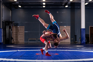 Two strong wrestlers in blue and red wrestling tights are wrestlng and making a  making a hip throw  on a yellow wrestling carpet in the gym. Young man doing grapple. - obrazy, fototapety, plakaty