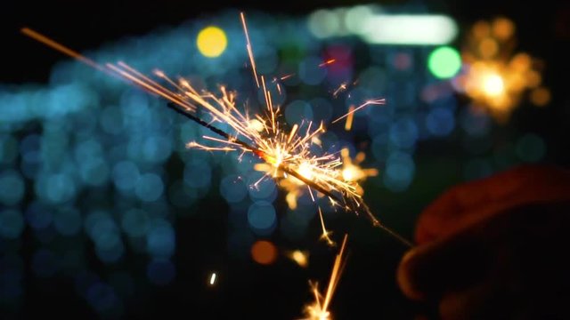 Hand of young woman holding a sparkler burning with lights bokeh in background, slow motion shot.
