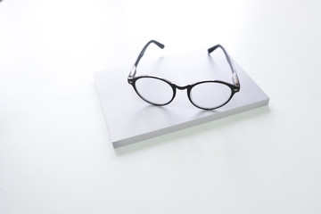 Black eye glasses are placed on a stack of books on a white wooden table beside the window.