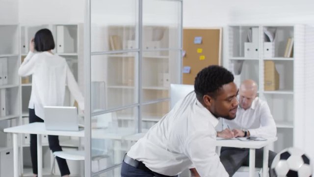 Thigh-up shot of young African American office worker having fun break with football in open space office, bouncing it with knee and catching, while colleagues are busy working behind computers