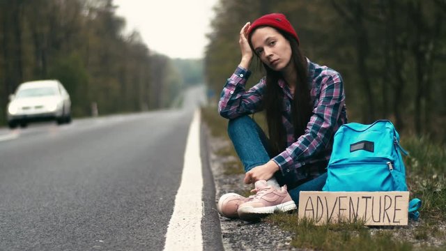 Lonely girl sitting at roadside waiting for car. Female hitchhiker sitting bored at side of local empty road slow motion 4k portrait. Difficulties of travelling alone danger wildness hipster concept