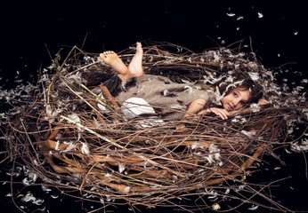 girl with long brown hair in a linen dress with feathers ,lying bent legs,in a decorative nest of branches with decorative white eggs.