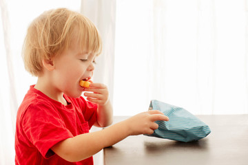 Side view of blonde child in red t-shirt with pleasure eating corn sticks holding and looking in...