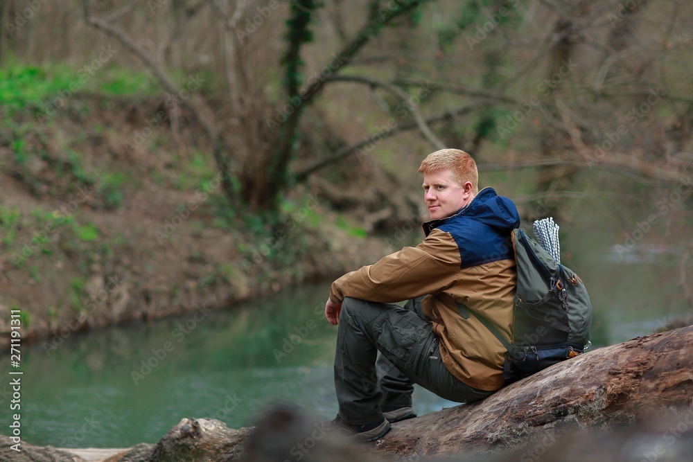 Wall mural redhead man in a brown-and-blue windbreaker with a backpack sits on a log by the river in the spring