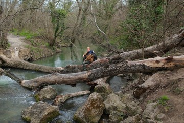 Redhead man in a brown-and-blue windbreaker with a backpack sits on a log by the river in the spring in the Crimea. Travel, adventure and hiking concept.