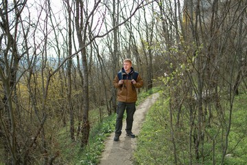 Obraz premium Redhead man in a brown-and-blue windbreaker with backpack stands on footpath in the woods in the Crimea in the early spring. Travel, adventure, hiking and motivation concept.