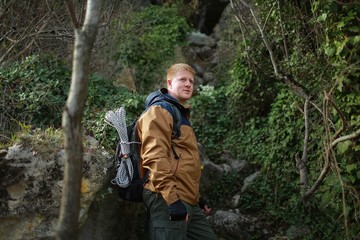 Naklejka premium Redhead man in a brown-and-blue windbreaker with backpack and rope stands in mountain forest in the Crimea in the early spring. Travel, adventure, hiking and motivation concept.