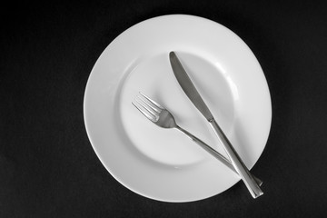 White empty clean plate and stainless knife and fork isolated on black background. Top view