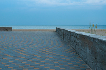 Chess pattern walkway near beach. Tranquil seascape in dusk with nobody and cement border between walkway and beach sand with high wild plants. Blurred moon in blue evening sky. Black sea landscape 