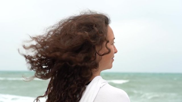Young woman on the background of the sea waving her head to the side and fluttering hair in the wind. Charming brunette girl on sea coast. Beautiful woman enjoys a wind, the feeling of freedom