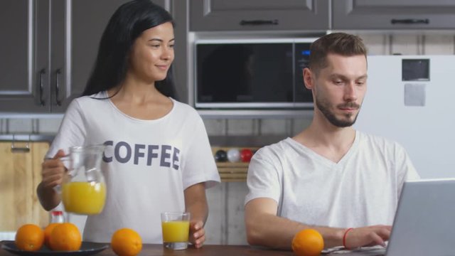 Young cheerful wife pouring orange juice and giving glass to husband while he working on laptop at kitchen table