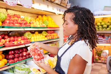 Serious young Black woman buying package of fresh organic strawberries in supermarket