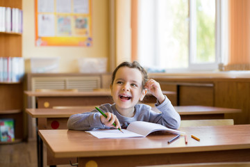 Caucasian little smiling girl sitting at desk in class room and begins to carefully draw in a pure notebook. Happy pupil