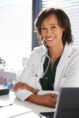 Portrait Of Smiling Female Doctor Wearing White Coat With Stethoscope Sitting Behind Desk In Office
