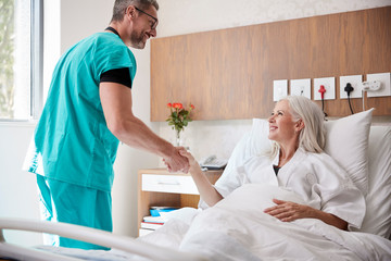 Surgeon Visiting And Shaking Hands With Mature Female Patient In Hospital Bed