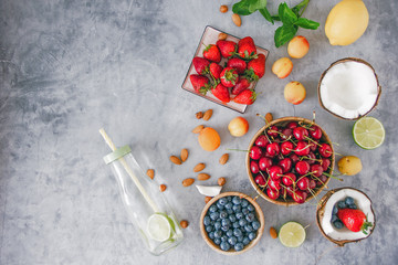 Top view Clear-colored cooling drink in a glass bottle with a straw, a number of broken coconut, strawberries, cherries, apricots, blueberries, lime, lemon, nuts on a dark stone background, save space