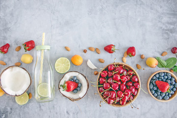 Top view Clear-colored cooling drink in a glass bottle with a straw, a number of broken coconut, strawberries, cherries, apricots, blueberries, lime, lemon, nuts on a dark stone background, save space