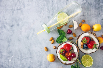 Top view Clear-colored cooling drink in a glass bottle with a straw, a number of broken coconut, strawberries, cherries, apricots, blueberries, lime, lemon, nuts on a dark stone background, save space