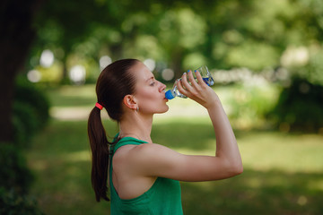 Portrait of young beautiful dark-haired woman wearing green t-shirt drinking water at summer green...