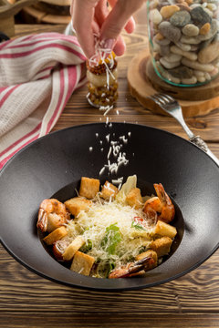 Female Hand Sprinkling Grated Parmesan Cheese On Fresh Vegetable Salad With Shrimp And Croutons