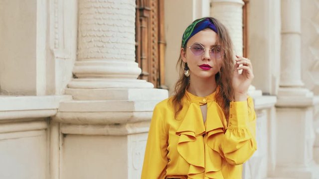 Street fashion portrait of young beautiful elegant lady wearing trendy purple color sunglasses, headband, stylish yellow blouse with jabot, posing in old european city
