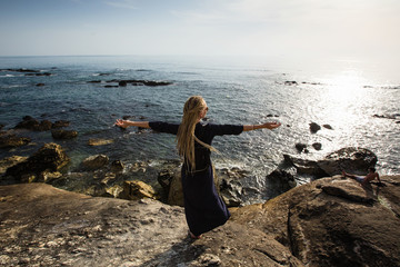 A young woman with long light dreadlocks stands on the rocky shore of the ocean watching the surf.