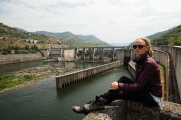 Fototapeta premium Young woman with dreadlocks sitting near a power plant in the mountains. Abandoned industrial facilities.