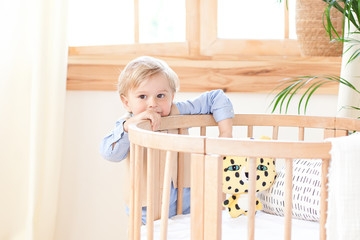 A boy stands alone beside a cot in the nursery. Lonely baby is in kindergarten near the crib. Loneliness. Eco-friendly children's room decor in the Scandinavian style.  The boy is at home.  interior