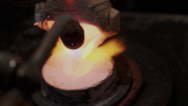 Molten gold, platinum or silver pouring into shape form while heating with gasoline burner. Usual work process of professional jeweler or goldsmith. Liquid precious metal. Closeup, slow motion.