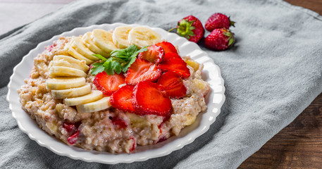 Oatmeal porridge with milk, strawberry, banana in white plate on wooden table background