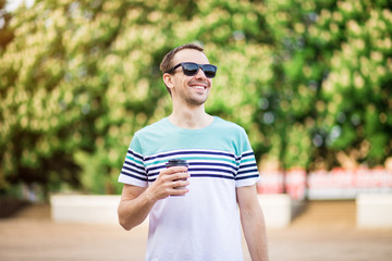 Fototapeta na wymiar Attractive young man with dark hair wearing sunglasses is walking the street with a cup of coffee on a sunny day. Bearded hipster guy wearing casual clothes enjoys relaxing on a summer weekend.