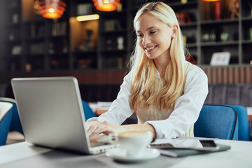 Fototapeta premium Beautiful smiling Caucasian blonde female blogger dressed smart casual sitting in cafeteria and using laptop.