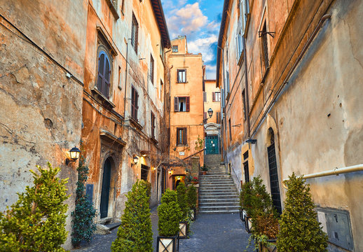 Rome, Italy. Yard of old street in downtown with antique building and stone stairs. Evening cityscape with street lamps on walls and decorative plants in flowerpots.