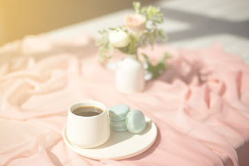 Fototapeta na wymiar French macaroon blue plate on the pink and pink coffee cup standing on a wooden table with a pink tablecloth white vase with flowers roses and greens