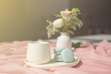 French macaroon blue plate on the pink and pink coffee cup standing on a wooden table with a pink tablecloth white vase with flowers roses and greens